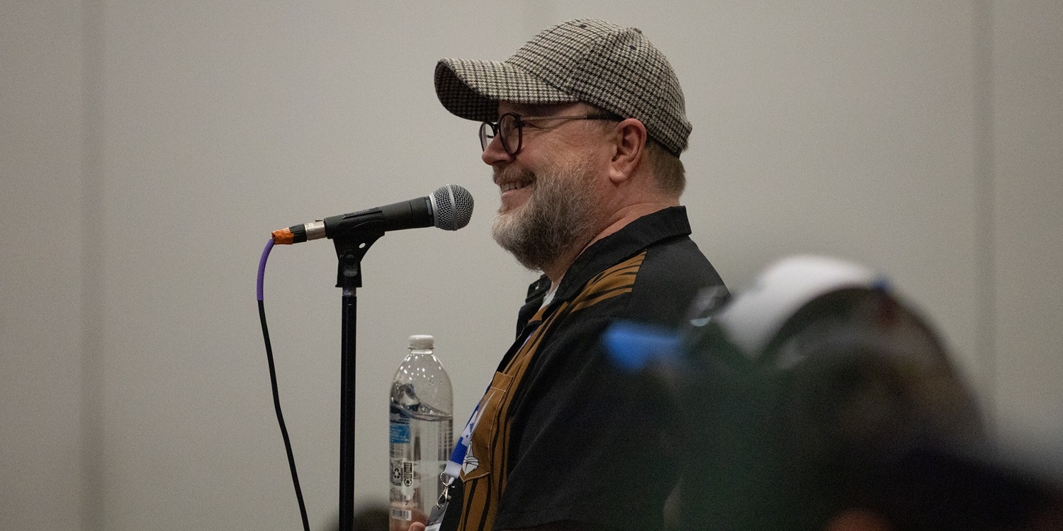 A smiling man speaks into a microphone during a panel or live event. He is shown in profile, wearing a patterned flat cap, round glasses, and a dark jacket with brown accents. A microphone on a stand is positioned close to his mouth, with a purple cable visible. A clear water bottle sits on the table in front of him. The background is a plain, light-colored wall, and the foreground includes a slightly blurred audience member, emphasizing the speaker as he addresses the room.
