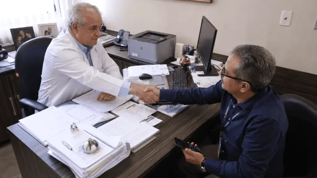 Médico de jaleco branco aperta a mão de um paciente ou colega de trabalho sentado em frente à sua mesa em um consultório. A mesa tem papéis acumulados, um computador e uma impressora. Imagem sobre o uso de LLMs e inteligência artificial no letramento digital em saúde.