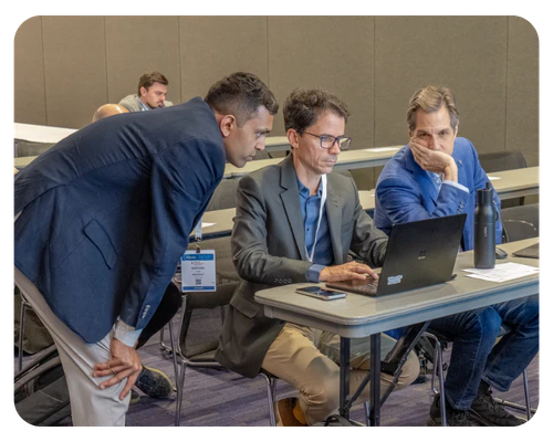 Three men seated at a table, using a laptop, engaged in discussion or collaboration.