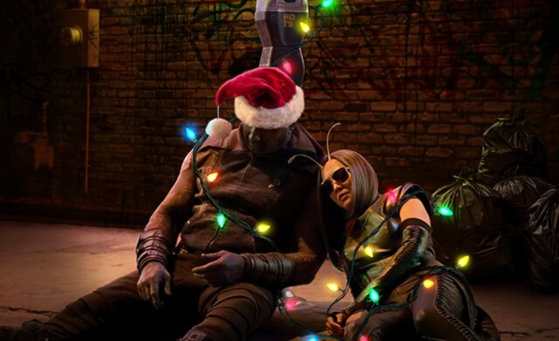 Drax and Mantis sit on a sidewalk, leaning on a post in front of garbage bags. Drax is slumped over, wearing a Santa hat, Mantis is wearing sunglasses--the pair are wrapping in lights.