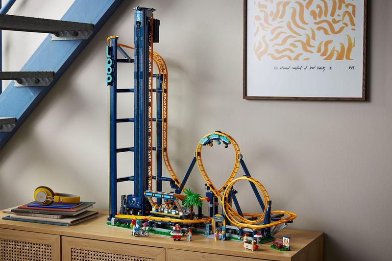 Loop Coaster Lego Set with a large drop and two loops. There is one car going right now. The set is on a cabinet with a stack of thin books and yellow over the ear head phones. It is under a blue staircase and there is a framed patterned print.