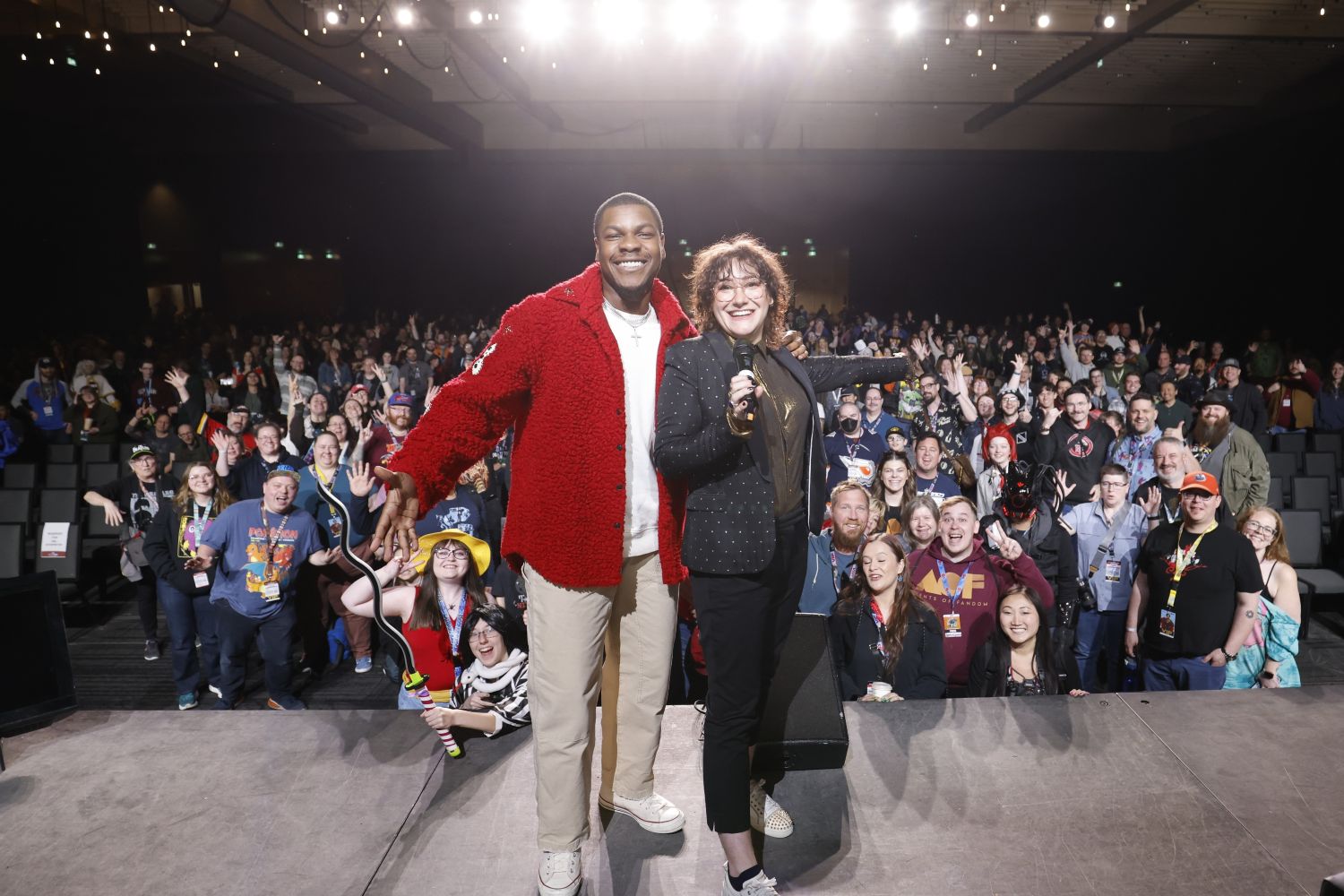 Actor John Boyega and a FAN EXPO host stand on stage in front of a cheering crowd during a spotlight panel. Boyega wears a red textured jacket and both are smiling with arms outstretched as fans wave in the background.
