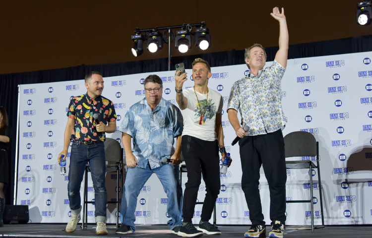 From left to right, Elijah Wood, Sean Astin, Dominic Monaghan, and Billy Boyd standing on a stage waving to the crowd