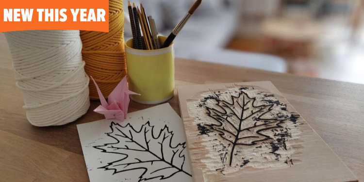 A wooden table with a cozy craft setup displays macrame rope, paintbrushes, and a finished block print of a maple leaf. A pink origami crane sits beside the crafting supplies.