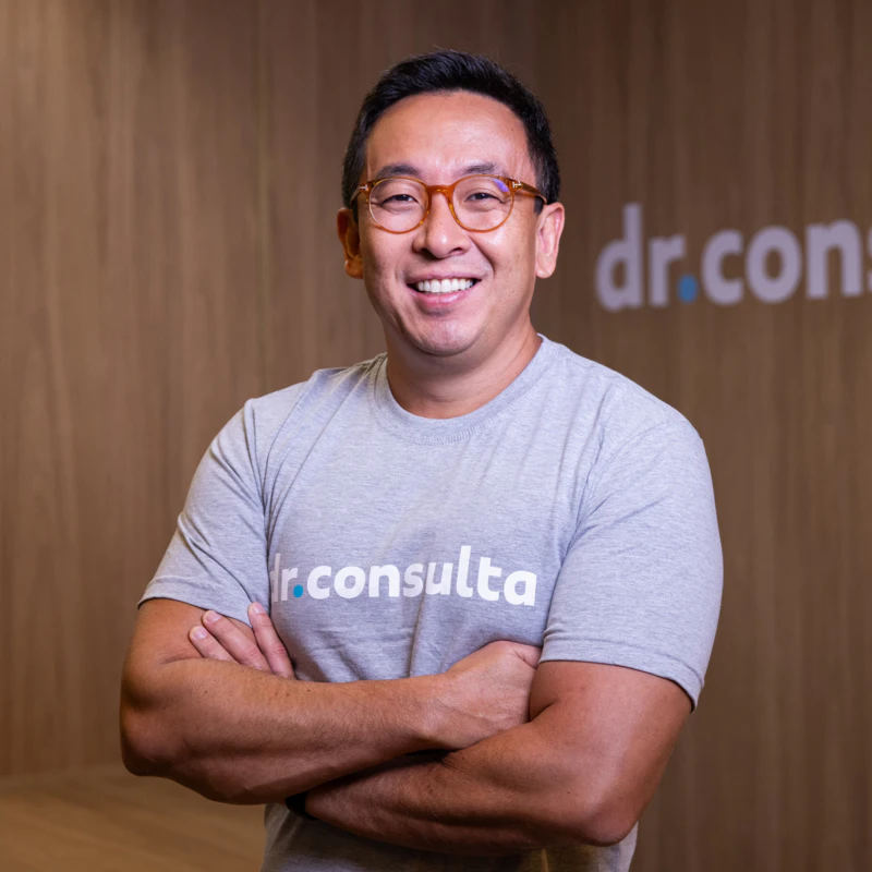 Massanori Shibata Jr., CEO da dr.consulta, sorrindo com braços cruzados vestindo camiseta cinza da marca e óculos de armação clara.