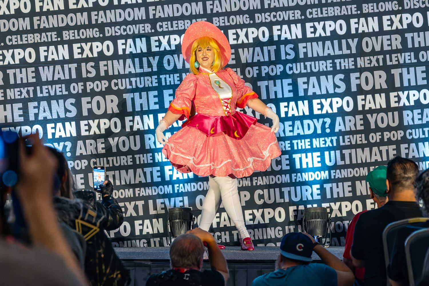 A wide shot of the competition stage shows a cosplayer in a pink frilly costume performing for the audience, with attendees in the foreground photographing the moment.