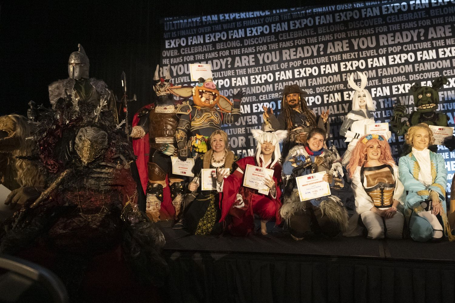 A large group of cosplayers from the competition pose together on stage, holding certificates and dressed in a variety of elaborate costumes, from armored warriors to fantasy characters.