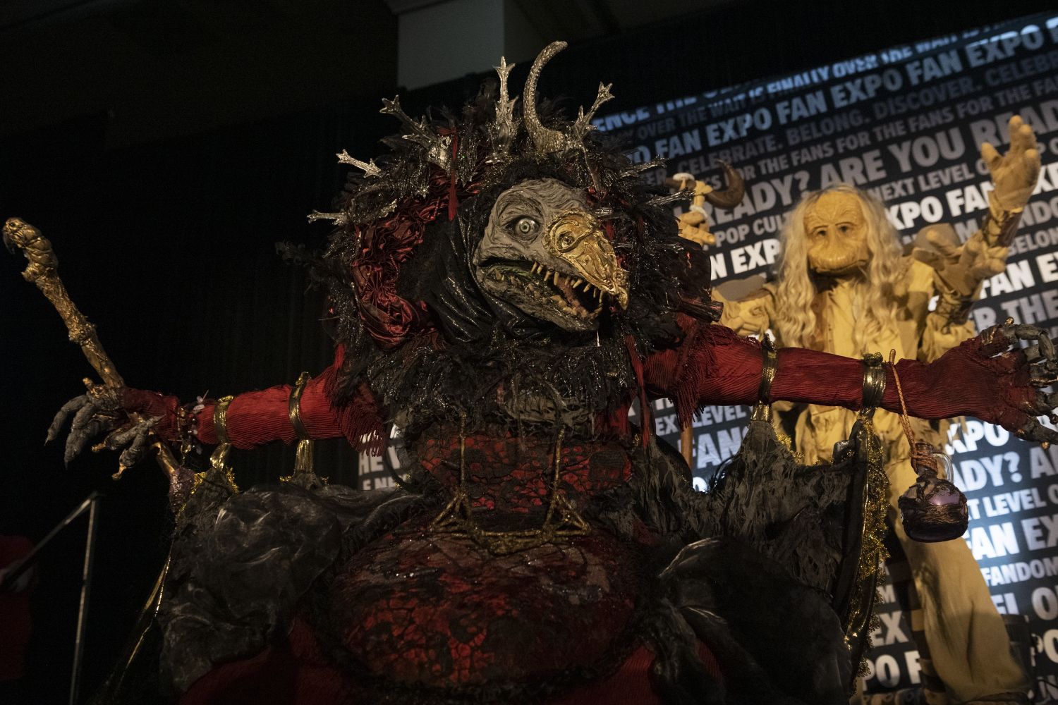 Two elaborate Dark Crystal cosplays stand on stage: a Skeksis in a detailed black-and-red costume with outstretched arms and a Mystic with long white hair and tan robes behind them.
