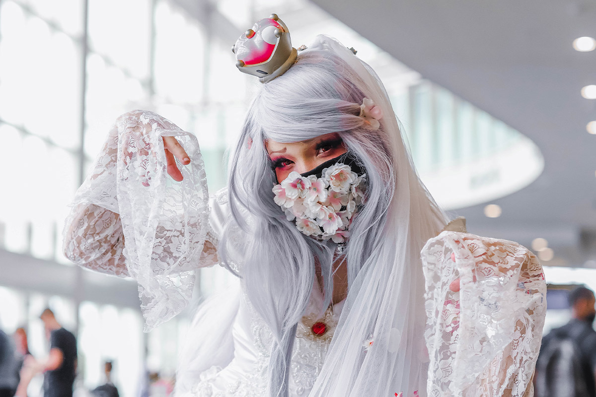 A cosplayer with a light grey wig, floral white lace arm sleeves and a small white & pink crown poses with her hand extended above her