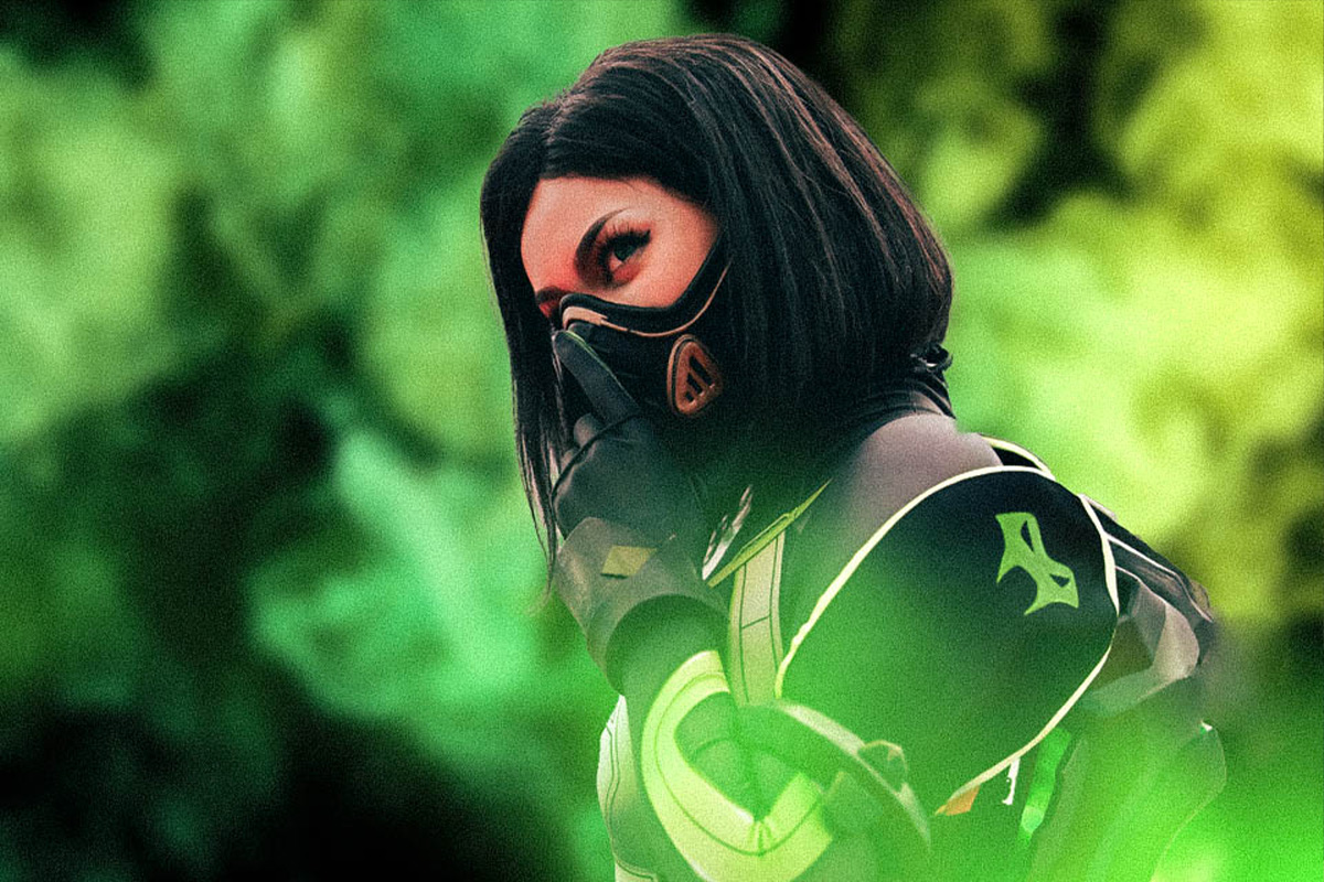 A cosplayer dressed in a black wig with a black mask and black & gold armour poses with a green background and green mist around her