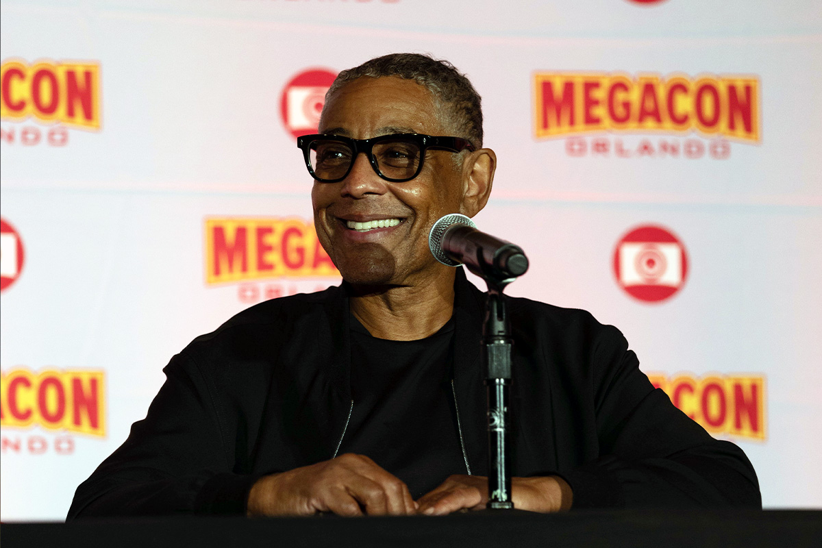 Giancarlo Esposito on stage sitting in front of a microphone smiling while wearing a black jacket and black glasses