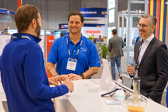 Three men engage in conversation at a bustling trade show, surrounded by various booths and attendees