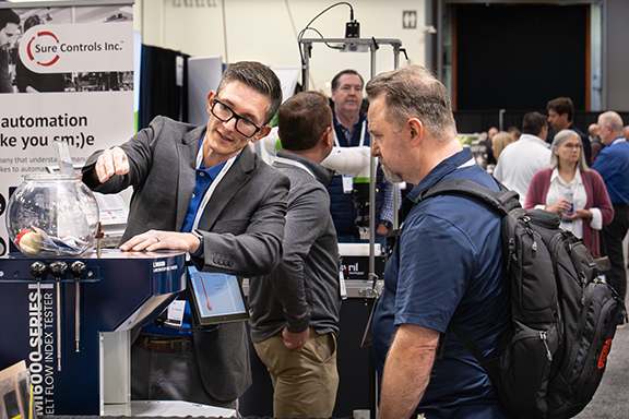 A man stands beside a machine at a trade show, engaging with attendees and showcasing the equipment's features