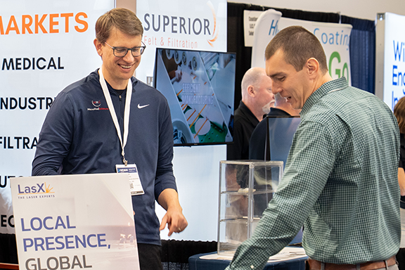 Two men stand at a trade show table, engaging with attendees beside a prominently displayed sign