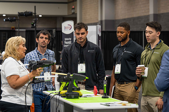 A group of people gathers around a table as a man demonstrates a tool he is holding
