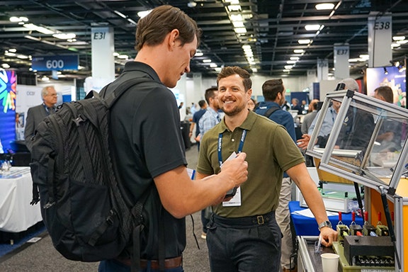 wo men engaged in conversation at a convention, surrounded by attendees and displays in a bustling environment