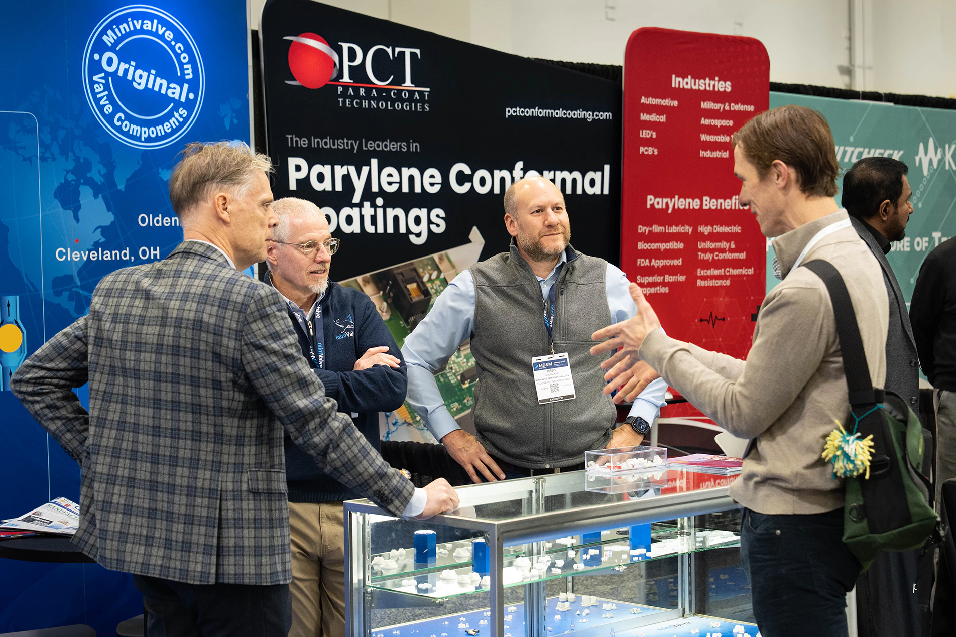 Four men converse at a trade show booth for Parylene conformal coatings, with informational banners and display cases visible.