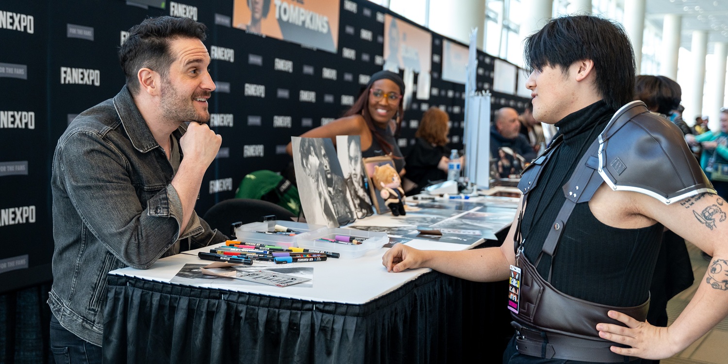 Nick Apostolides smiles and chats with a fan across an autograph table covered in markers and photo prints; the fan wears dark leather shoulder armor and a show badge, with FAN EXPO backdrop banners behind them.