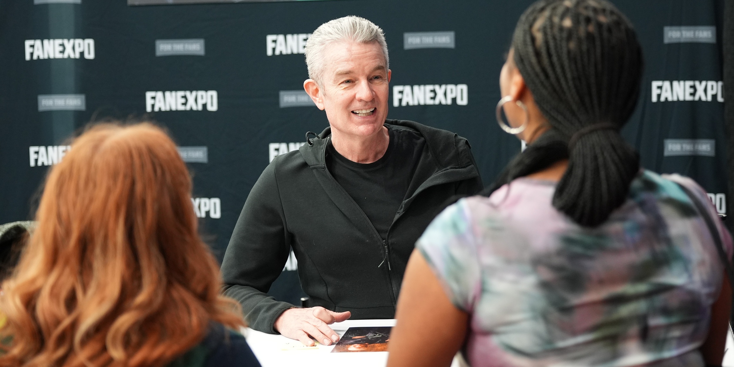 James Marsters sits at an autograph table, speaking with two fans and gesturing as he smiles, with a black FAN EXPO–branded backdrop behind him.