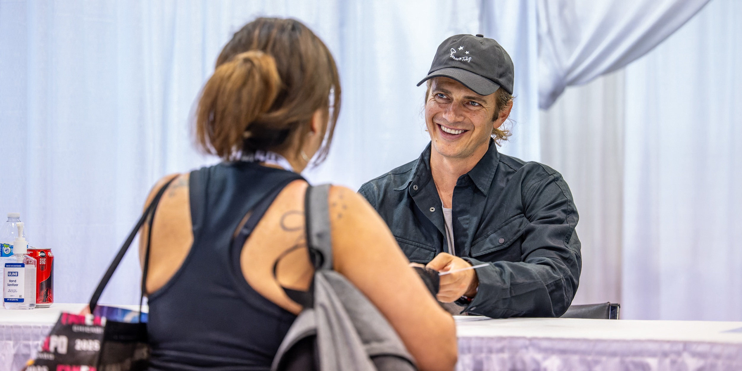 Hayden Christensen smiles while handing an autograph to a fan seated across a signing table at FAN EXPO Cleveland, with white draping behind them and personal items visible on the table.
