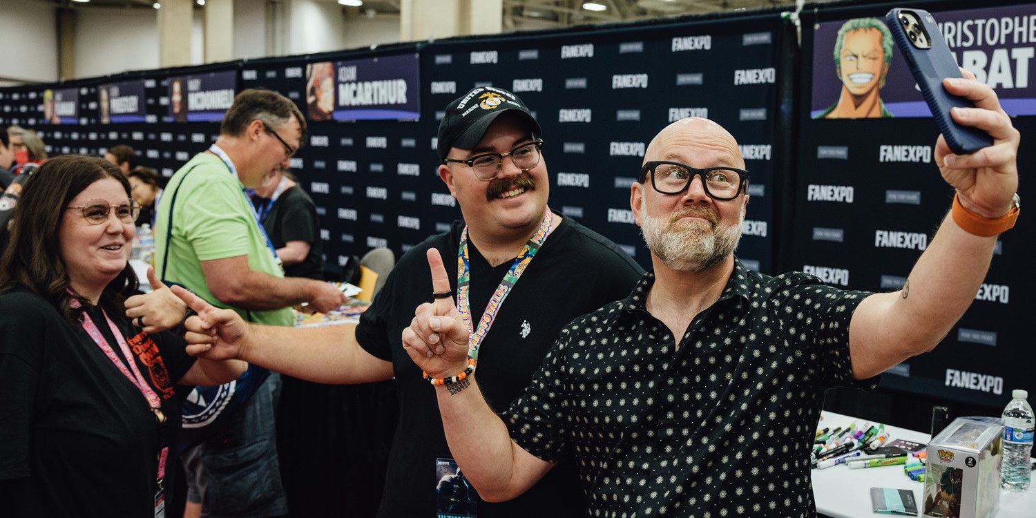 A group of excited anime fans gathers around voice actor Christopher Sabat, best known for roles like Vegeta in Dragon Ball Z and All Might in My Hero Academia, to take a selfie at his autograph table. Sabat smiles warmly and leans in with the group, holding up a peace sign. Fans are beaming, some wearing anime-themed T-shirts and cosplay accessories. Behind them, the table is covered with autograph prints and character art. The atmosphere is joyful and intimate, capturing a cherished moment of connection between fans and a beloved anime icon.