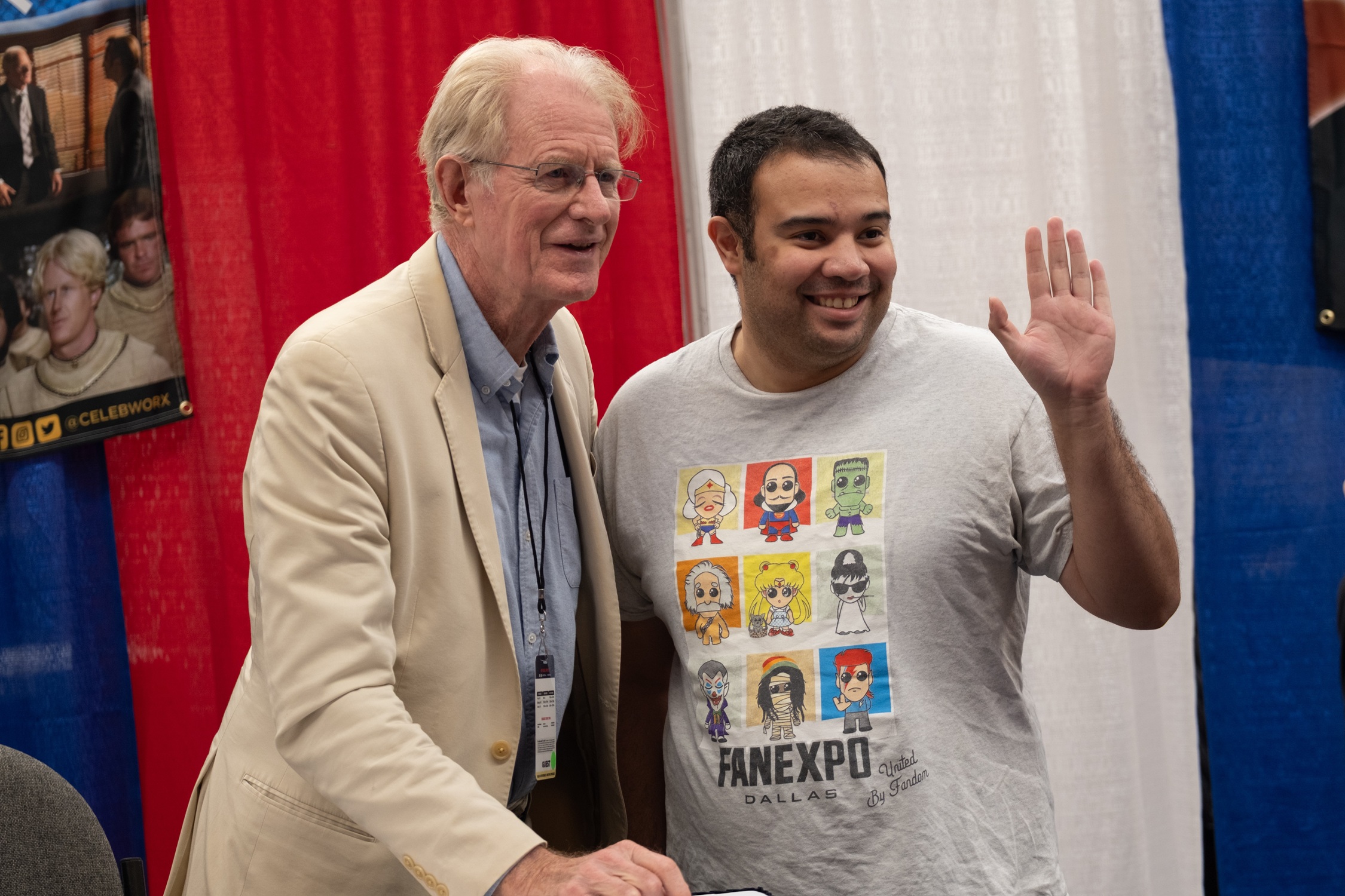 Actor Ed Begley Jr. smiles beside a fan wearing a FAN EXPO Dallas T-shirt featuring chibi superheroes and villains as they pose for a photo at his autograph table.