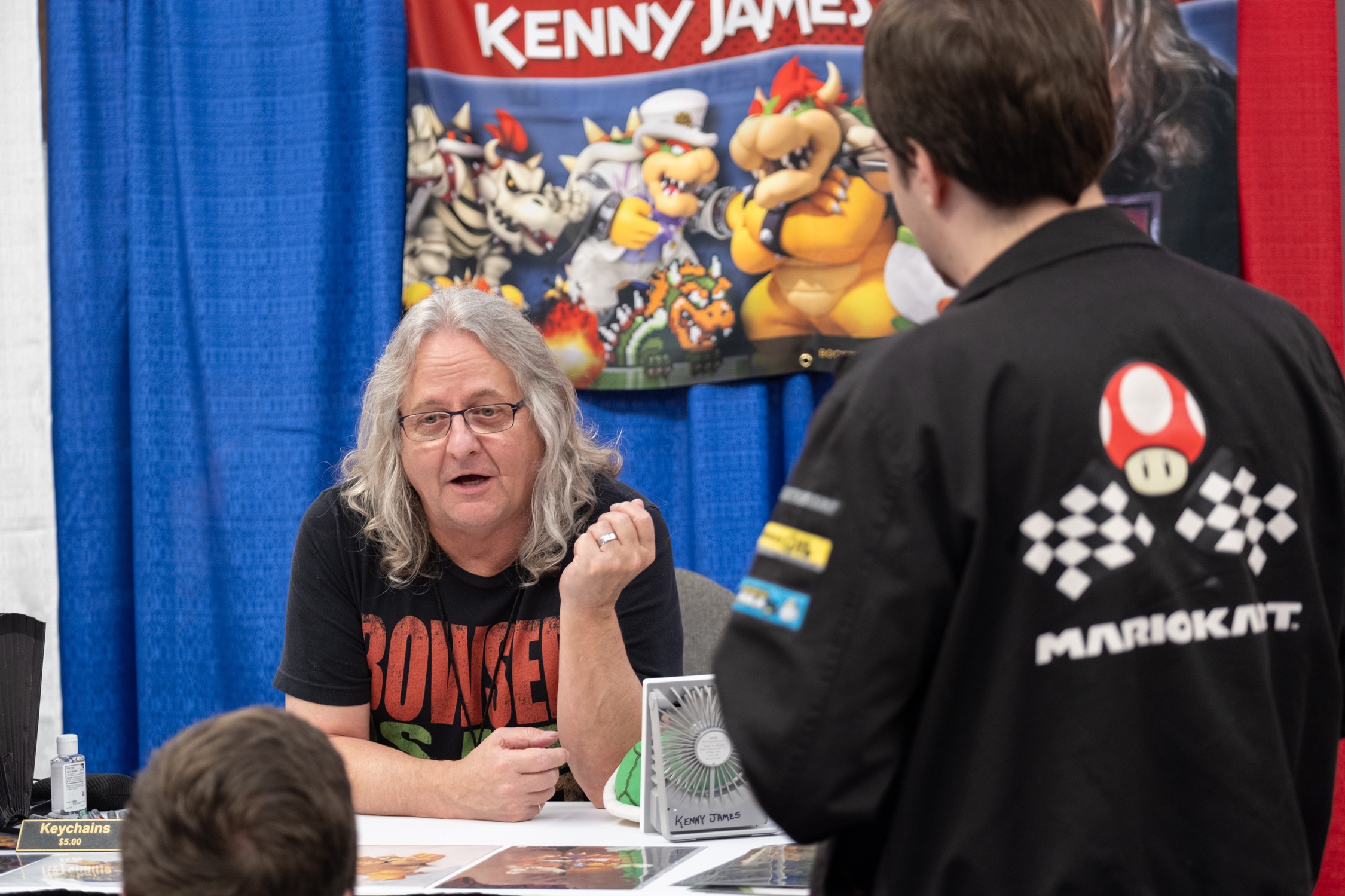 Voice actor Kenny James chats with fans at his autograph table, wearing a Bowser-themed shirt with a colorful banner of Mario characters behind him.