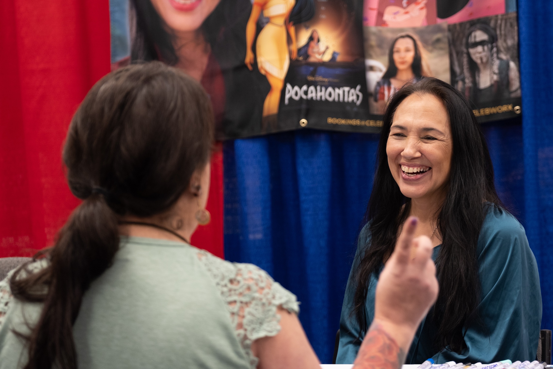 Actor Irene Bedard laughs warmly while meeting a fan at her table. Behind her, a banner highlights her animated and live-action roles, including Disney’s Pocahontas.