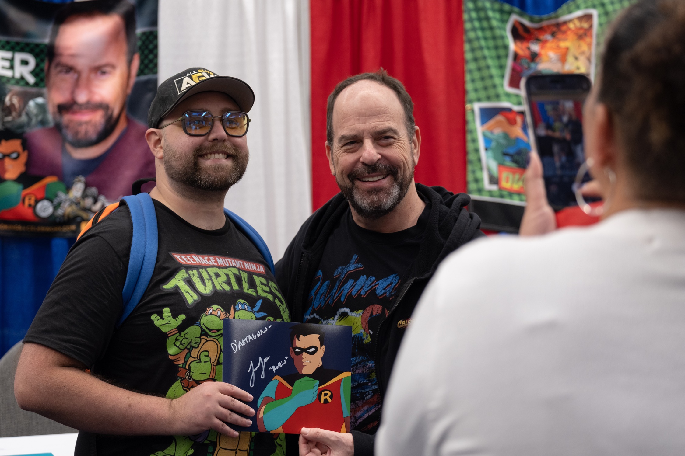 Voice actor Loren Lester poses with a fan holding a signed print of Robin from Batman: The Animated Series while another guest takes their photo in front of his booth banner.