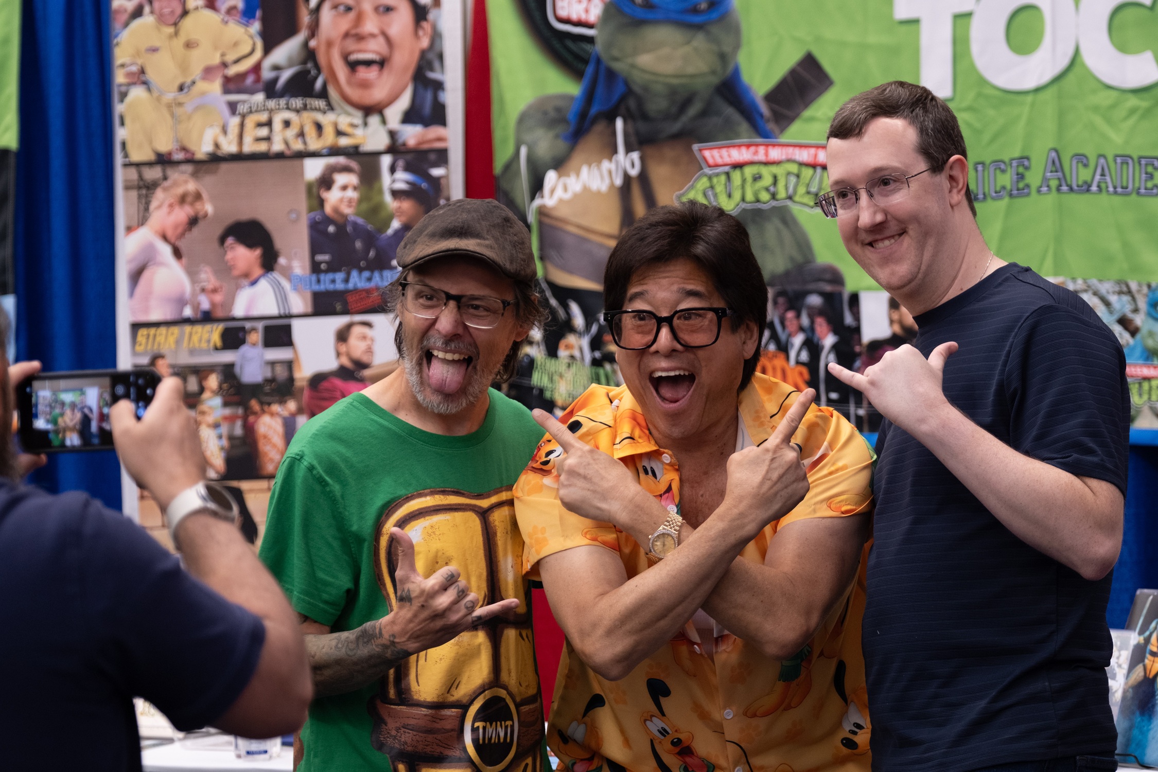Actor Brian Tochi enthusiastically poses with two fans at his booth, all smiling and gesturing playfully in front of a banner featuring Teenage Mutant Ninja Turtles and other classic roles.