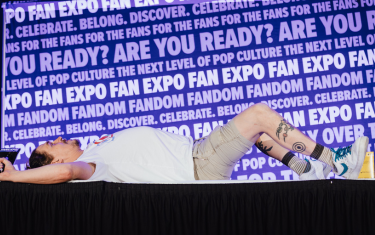 Jason Mewes laying on top of a table, with his arms above his head and his knees spread apart. He's wearing a white t-shirt, beige shorts, and white and blue sneakers.