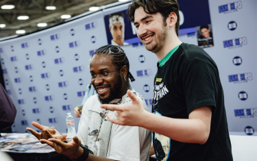 Shameik Moore smiling and posing with a fan while doing the Spider-Man web slinging pose. Shameik is on the left, wearing a white t-shirt, and the fan on the right is wearing a black Spider-Man t-shirt.