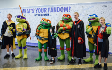 Four cosplayers dressed as the Teenage Mutant Ninja Turtles, posing with four boys cosplaying as Harry Potter characters. They are standing in front of a banner labeled "What's Your Fandom? My Fandom Is..."