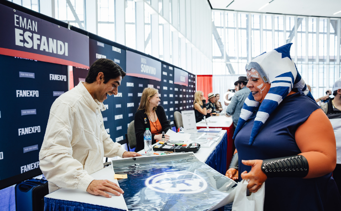 Eman Esfandi in a white shirt intently examines a large poster on a table at his booth. Beside him, a fan dressed in an Ahsoka cosplay, complete with face paint, helmet, and arm guard, excitedly interacts with him. In the background, other convention attendees and booths are visible, adding to the bustling atmosphere.