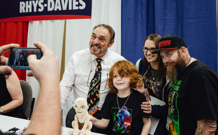 John Rhys-Davies in a white shirt and colorful tie shares a moment with a family at a convention booth. A young child with bright red hair and a graphic tee, a woman with glasses, and a bearded man wearing a cap are all posing for a photo being taken by another individual, as evidenced by the camera visible in the foreground. On the table is a Gollum doll.