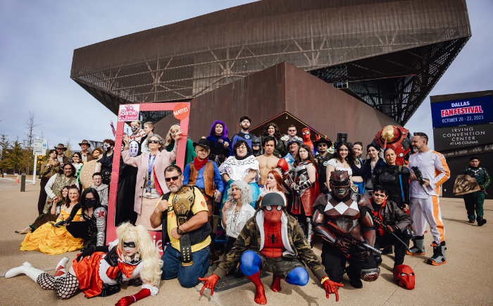 A large, diverse group of cosplayers gather outside the Irving Convention Center for a group photo. The participants are dressed as various fictional characters, including superheroes, villains, and fantasy figures. Costumes range from elaborate armors and flowing gowns to masks and vibrant wigs. The scene captures the festive spirit and creativity of the cosplay community.