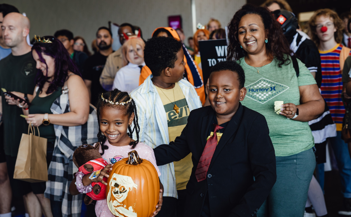 A vibrant gathering of people at Dallas FAN FESTIVAL. In the foreground, a cheerful young girl with braided hair holds a pumpkin with a painted face, accompanied by a toy doll. Beside her, a young boy smartly dressed in a suit and red tie interacts with an older boy in casual attire. A smiling woman, possibly their mother, stands proudly next to them, holding a white object. In the background, various attendees, some in costume, chat and socialize, with signs and banners slightly visible, indicating a lively, festive atmosphere.