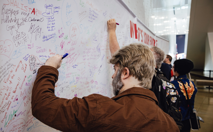 A man in a brown jacket, with a beard and swept-back hair, is signing on a large white wall filled with various handwritten messages, drawings, and signatures. Another individual stands beside him, reaching higher on the wall to add their own contribution. In the background, a few attendees walk by, one prominently dressed in a detailed blue jacket. The atmosphere is that of communal sharing and expression, with the wall acting as a collaborative canvas for attendees.