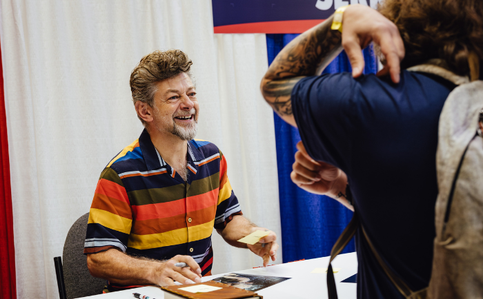 Andy Serkis, wearing a colorful striped shirt, sits at a table in a convention setting, happily engaging with a fan. As he signs an item, he shares a bright smile with the fan whose tattooed arm is in the foreground, capturing the moment with a camera.