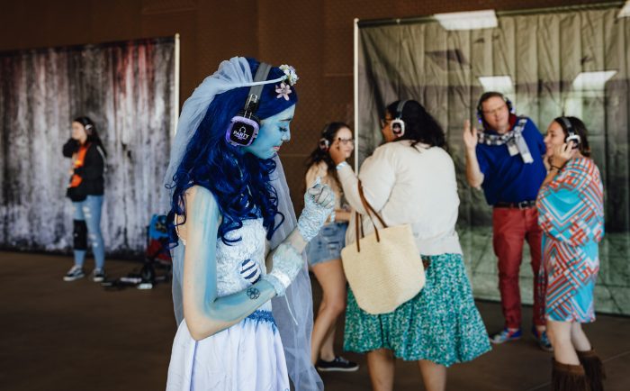 A woman cosplaying as Corpse Bride, with vivid blue skin, long blue hair, and ethereal white attire stands in the foreground, adorned with delicate flowers and headphones with a visible 'Radio' label. She is dancing in a silent disco. In the background, a few attendees also dance; notably, a man in a blue shirt gestures while talking to a woman in a vibrant patterned dress, and two younger women chat nearby. The setting is at a convention, with rustic backdrops set up for photo opportunities.