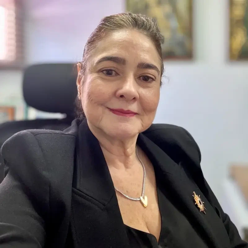 Mônica Bezerra sorrindo em seu escritório, vestindo blazer preto, blusa preta e colar com pingente de coração.
