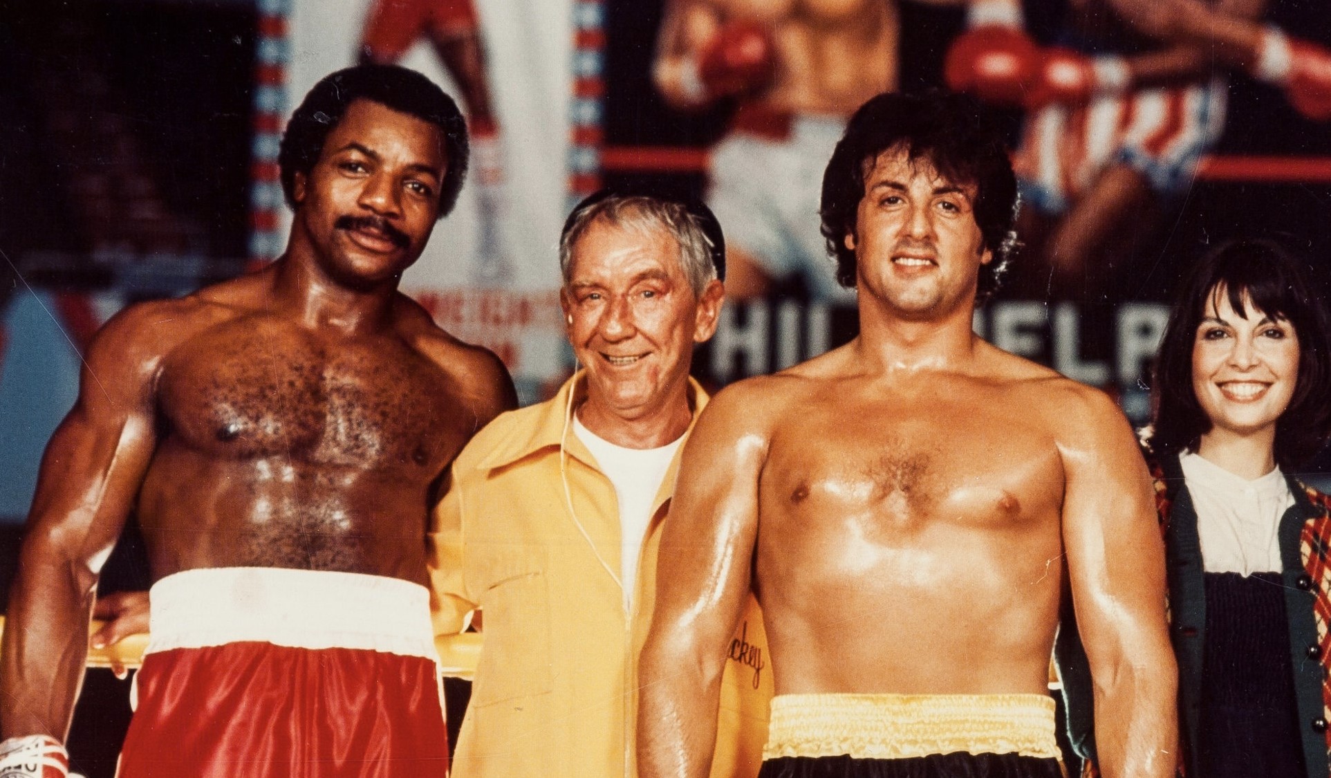 Carl Weathers, wearing red boxing shorts, standing next to Sylvester Stallone, wearing yellow and black shorts.