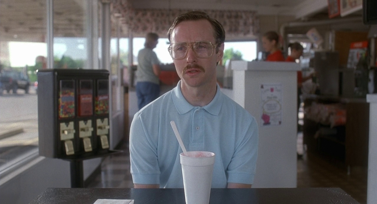 Kip sits in the diner with his strawberry milkshake in a Styrofoam cup. To his left, a candy machine with skittles, red hots, and runts, and behind him, patrons are talking with employees