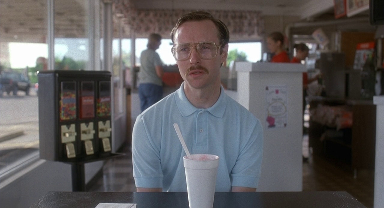 Kip sits in the diner with his strawberry milkshake in a Styrofoam cup. To his left, a candy machine with skittles, red hots, and runts, and behind him, patrons are talking with employees
