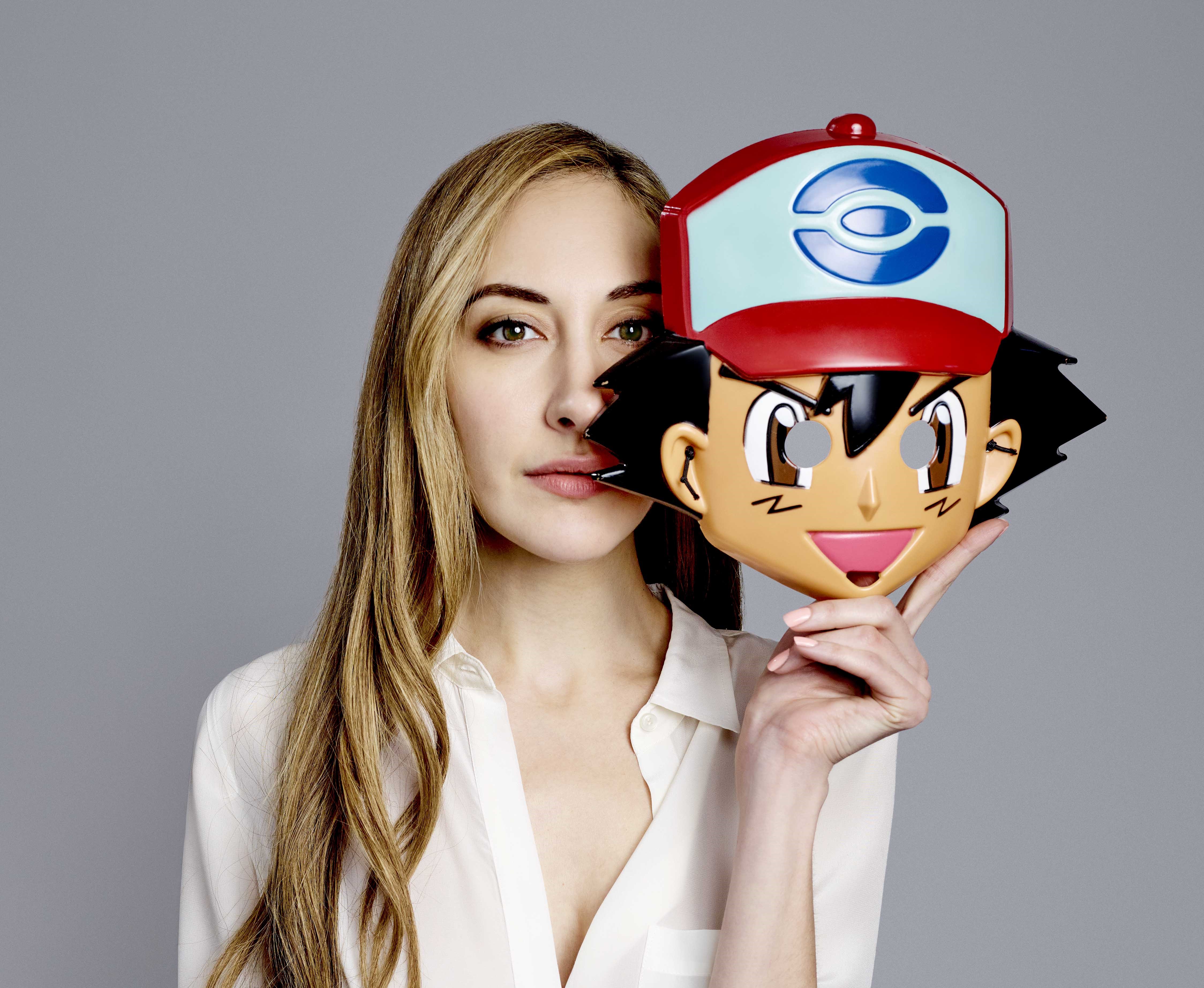 Sarah Natochenny in a white shirt holding an Ash Ketchum mask