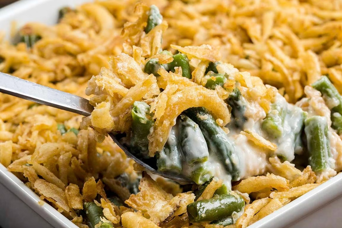 A green bean casserole sits on a table while a spoon takes a scoop of the food