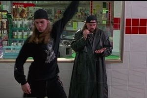Jay is wearing a black backwards cap, long sleeve shirt and pants. His left arm is up and his right arm is down as he is looking down. Behind him is Silent Bob who is wearing a black hat backwards and a long black coat. He has one hand by his face and the other hand is an open palm faced up. They are standing in front of a store window.