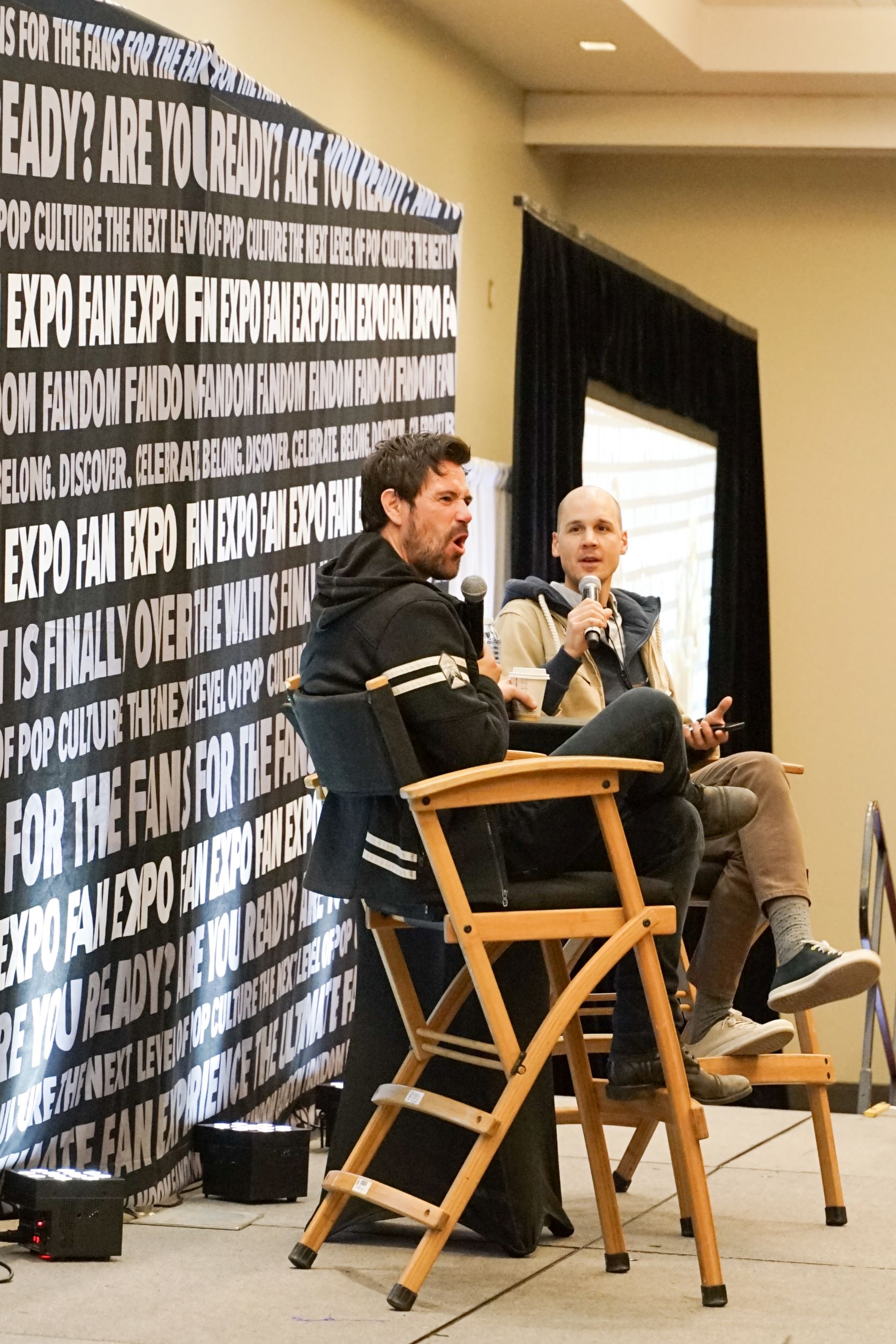 Matt Ryan sits on-stage at his panel at Toronto Comicon. He is wearing a black sweatshirt with white stripes on the sleeves.