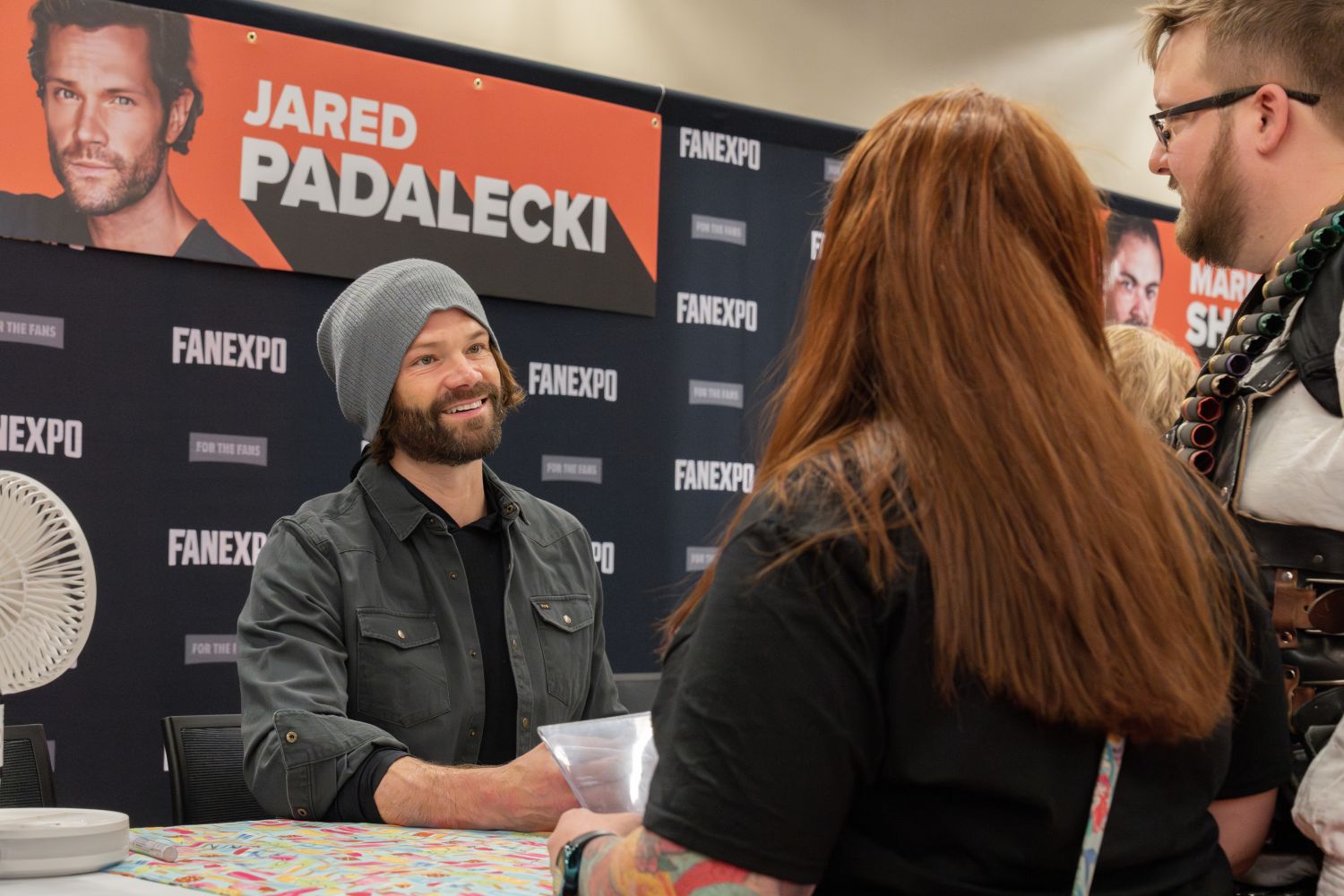 Jared Padalecki, wearing a gray beanie and dark button-up shirt over a black tee, smiles warmly while chatting with two fans at his table. One fan has long red hair and a black t-shirt, while the other, partially visible, wears glasses and a leather bandolier prop across his chest. Jared’s name is displayed in bold white text on an orange banner behind him, with the FAN EXPO logo on the background wall.
