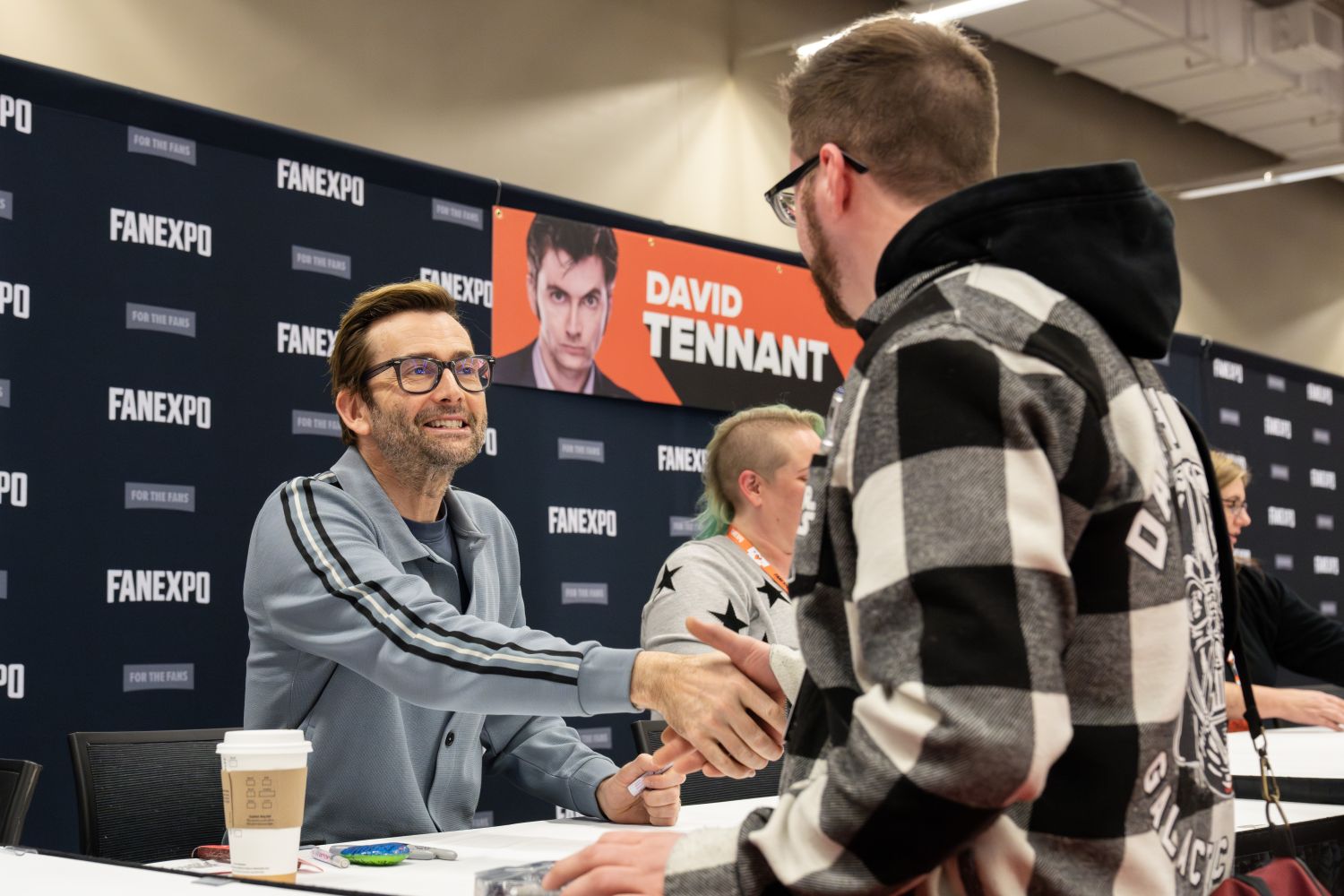 David Tennant, wearing a light blue zip-up jacket with dark stripes and glasses, enthusiastically shakes hands with a fan in a black-and-white plaid hoodie. David’s short hair is styled upward and he’s grinning widely. A Starbucks coffee cup and markers sit on his table, and behind him is a red and black banner with his name and a photo of him with a serious expression. Other attendees and staff can be seen in the background at neighboring autograph tables.